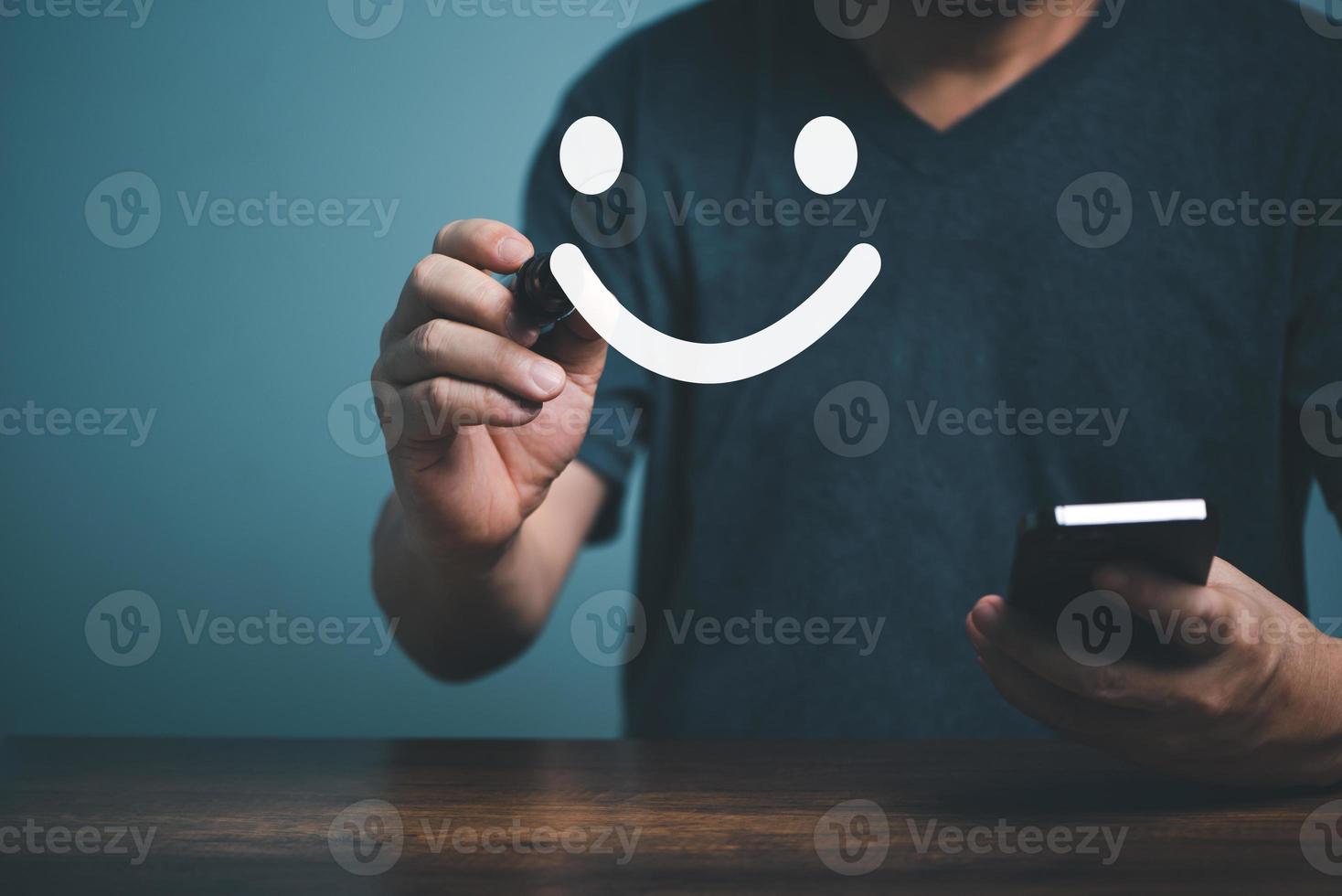 die hand des kunden zeigt ein feedback mit zufriedenem lächelndem gesichtszeichen und hält ein handy mit blauem hintergrund. servicebewertung, feedbackkonzept. foto