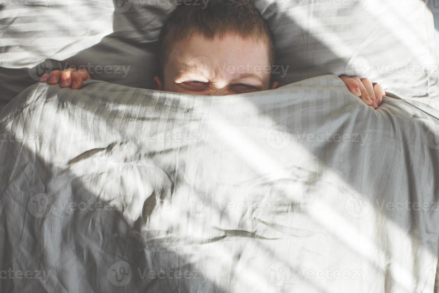 Junge schläft im Bett. Kind liegt auf Kissen und bedeckt sein Gesicht mit einer Decke foto