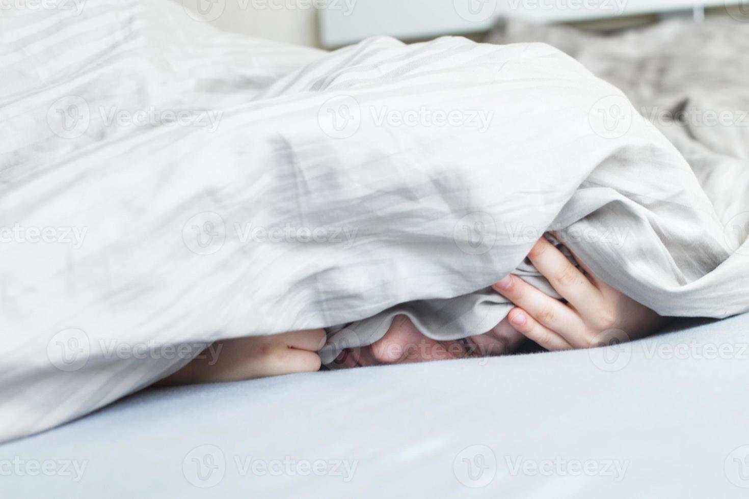 Junge morgens im Bett. Kind liegt und bedeckt sein Gesicht mit einer Decke foto