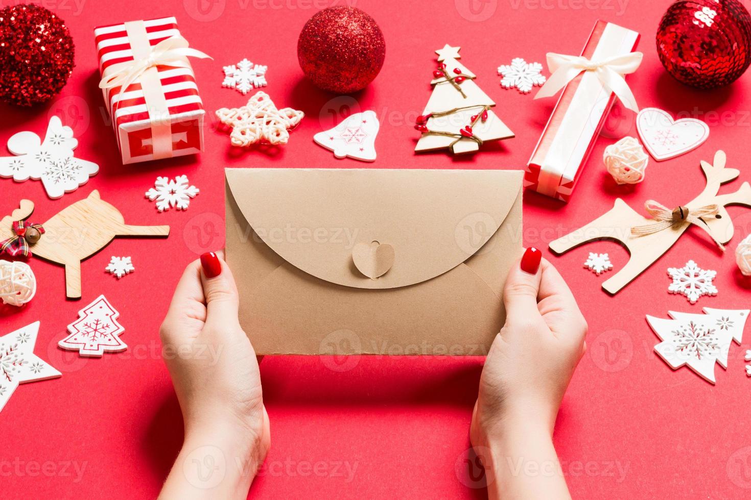 Draufsicht einer Frau, die einen Umschlag auf rotem Hintergrund aus Weihnachtsdekorationen hält. Weihnachtszeit-Konzept foto