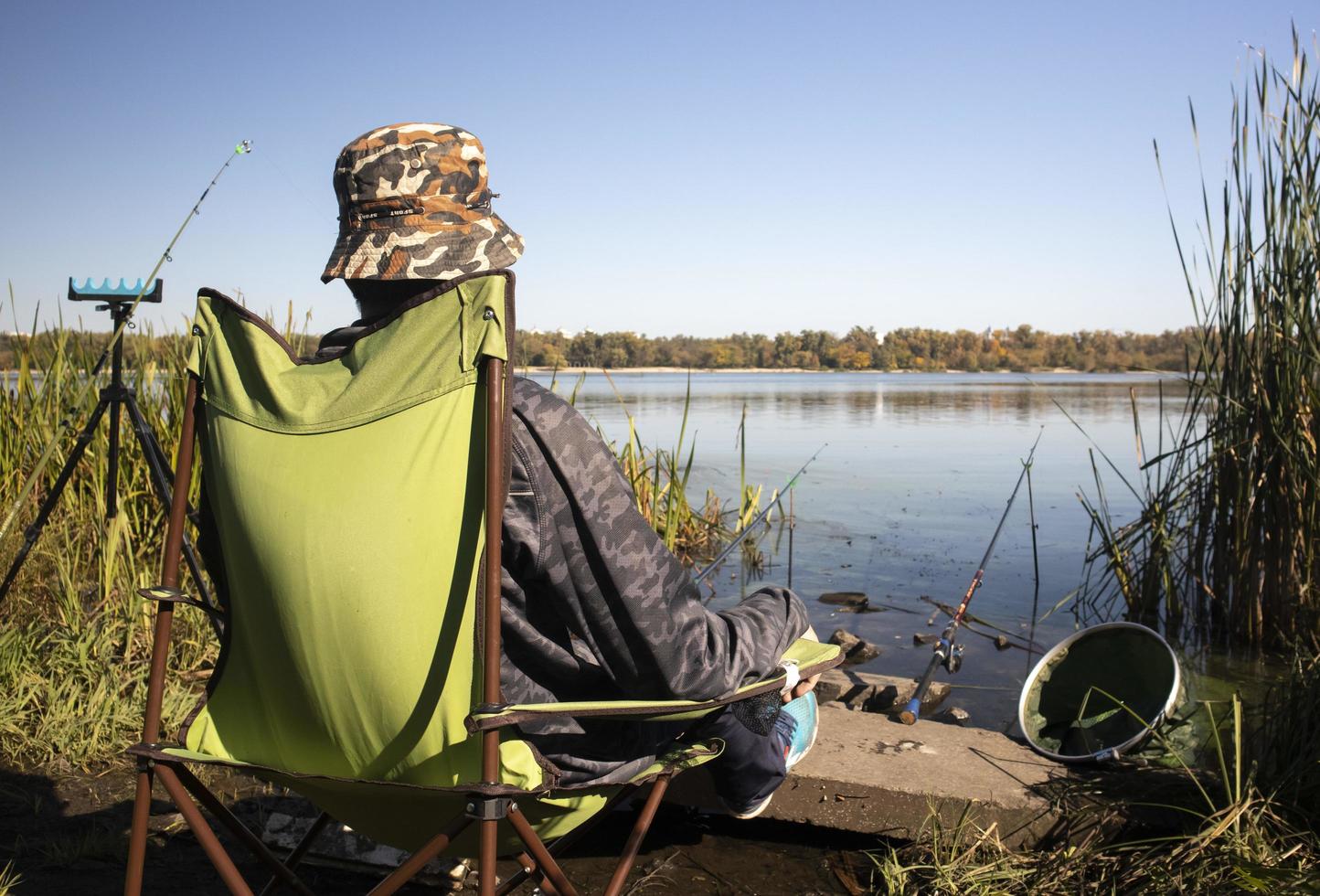 Der junge, nicht erkennbare Fischer fängt Fische, die auf einem Stuhl am Ufer eines Flusses oder Sees sitzen. Der Angler sitzt mit Angelausrüstung auf einem Stuhl. ein perfekter Urlaub am Fluss mit Angelrute. foto