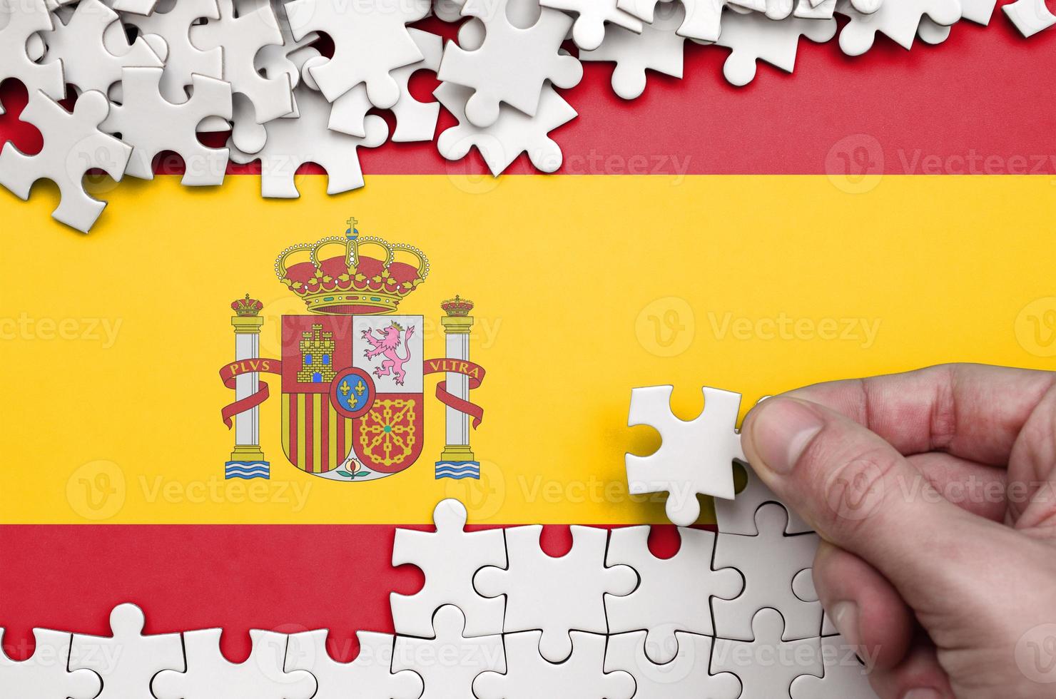 Die spanische Flagge ist auf einem Tisch abgebildet, auf dem die menschliche Hand ein weißes Puzzle faltet foto