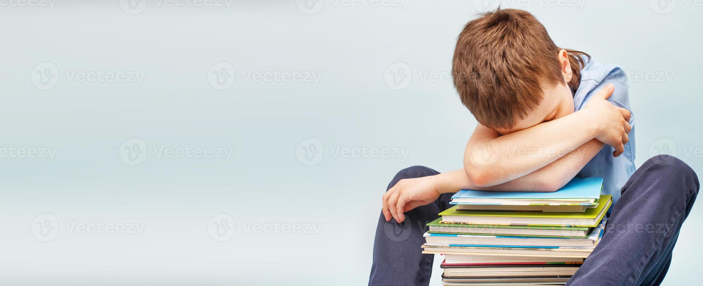 verärgerter Schuljunge, der mit einem Stapel Schulbücher sitzt und sein Gesicht mit den Händen bedeckt. Junge schläft auf einem Stapel Lehrbücher foto