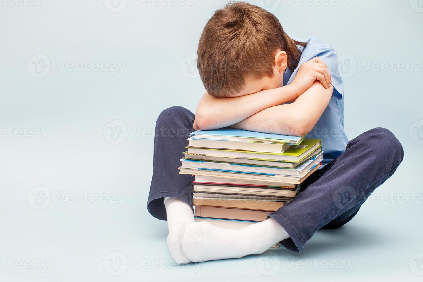 verärgerter Schuljunge, der mit einem Stapel Schulbücher sitzt und sein Gesicht mit den Händen bedeckt. Junge schläft auf einem Stapel Lehrbücher foto