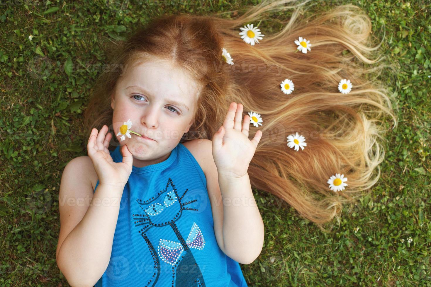 Porträt eines schönen kleinen Mädchens mit gesunden roten Haaren mit Kamillenblüten, die auf dem Gras liegen foto