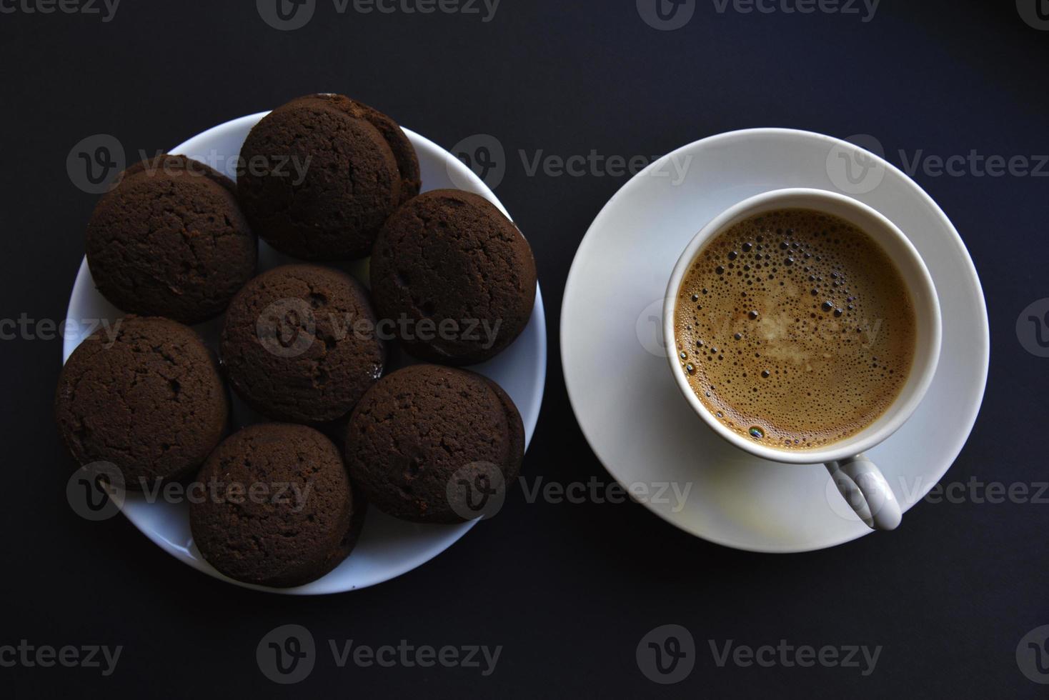 Köstlicher schwarzer Kaffee mit Schaum in einer weißen Tasse mit süßen Keksen. leckeres Frühstück mit Kaffee und Keksen. süße und weiche Kekse auf schwarzem Hintergrund. foto