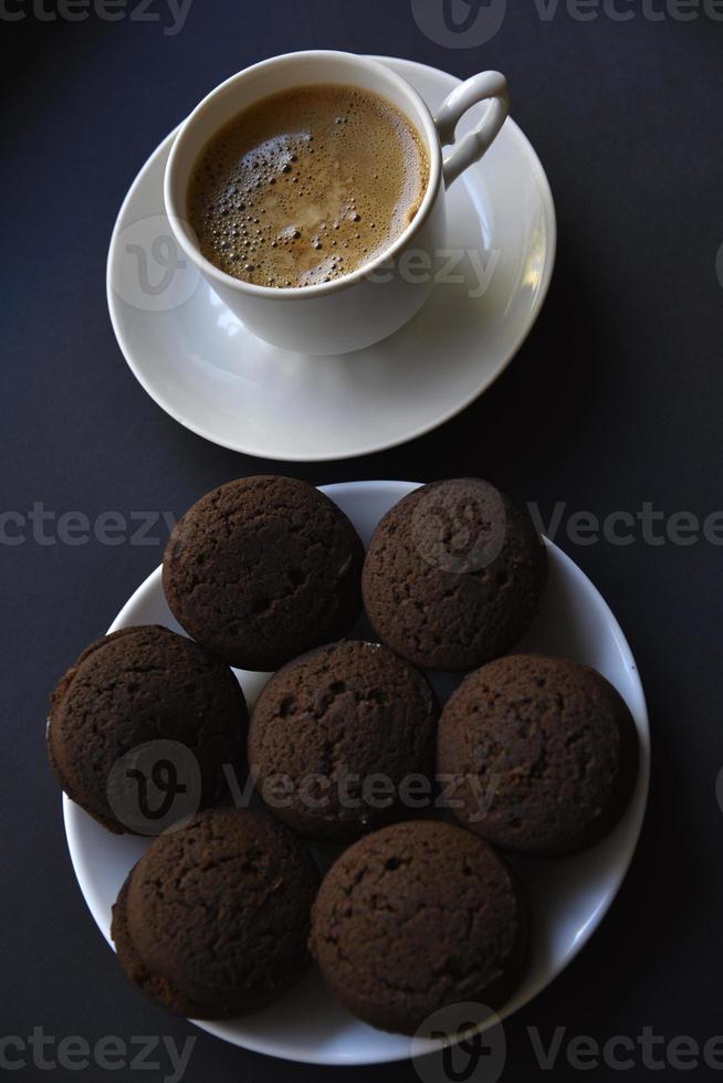 Köstlicher schwarzer Kaffee mit Schaum in einer weißen Tasse mit süßen Keksen. leckeres Frühstück mit Kaffee und Keksen. süße und weiche Kekse auf schwarzem Hintergrund. foto