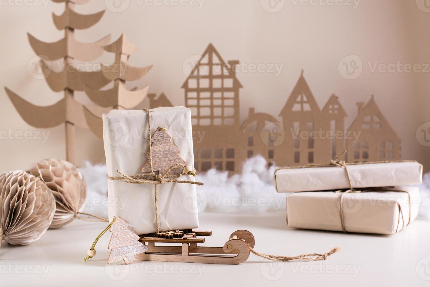 diy weihnachtliche wohndeko – geschenk aus bastelpapier auf einem schlitten, einem baum aus pappe und einem haus. handgefertigt foto