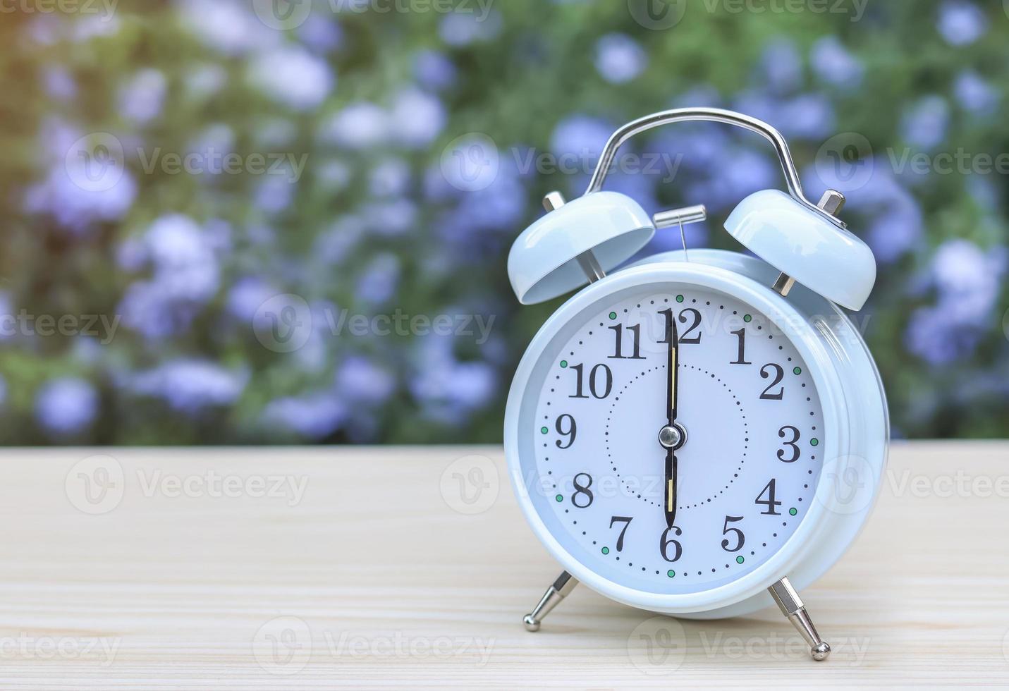 Vorderansicht des weißen Vintage-Weckers, der 6 Uhr auf dem Tisch im Garten mit violettem Blumenhintergrund zeigt. foto