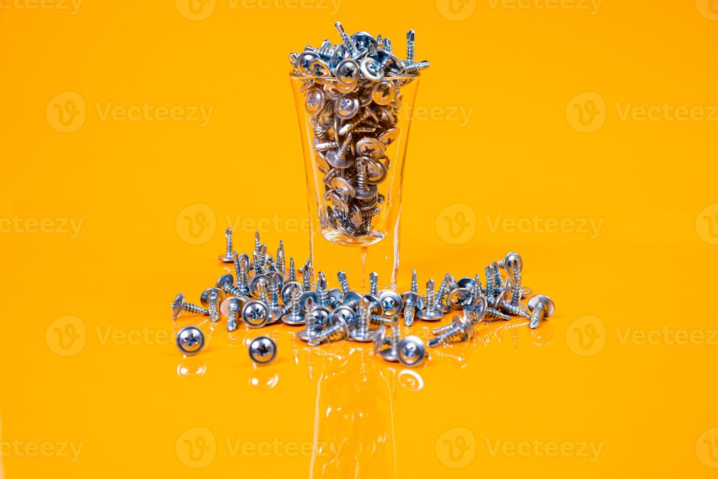 viele selbstschneidende Metallschrauben aus Stahl in einem Glas, in einem Eimerglas. selbstschneidende Schraube für Metall, für Eisen, verchromte selbstschneidende Schraube, auf orangegelbem Hintergrund, foto