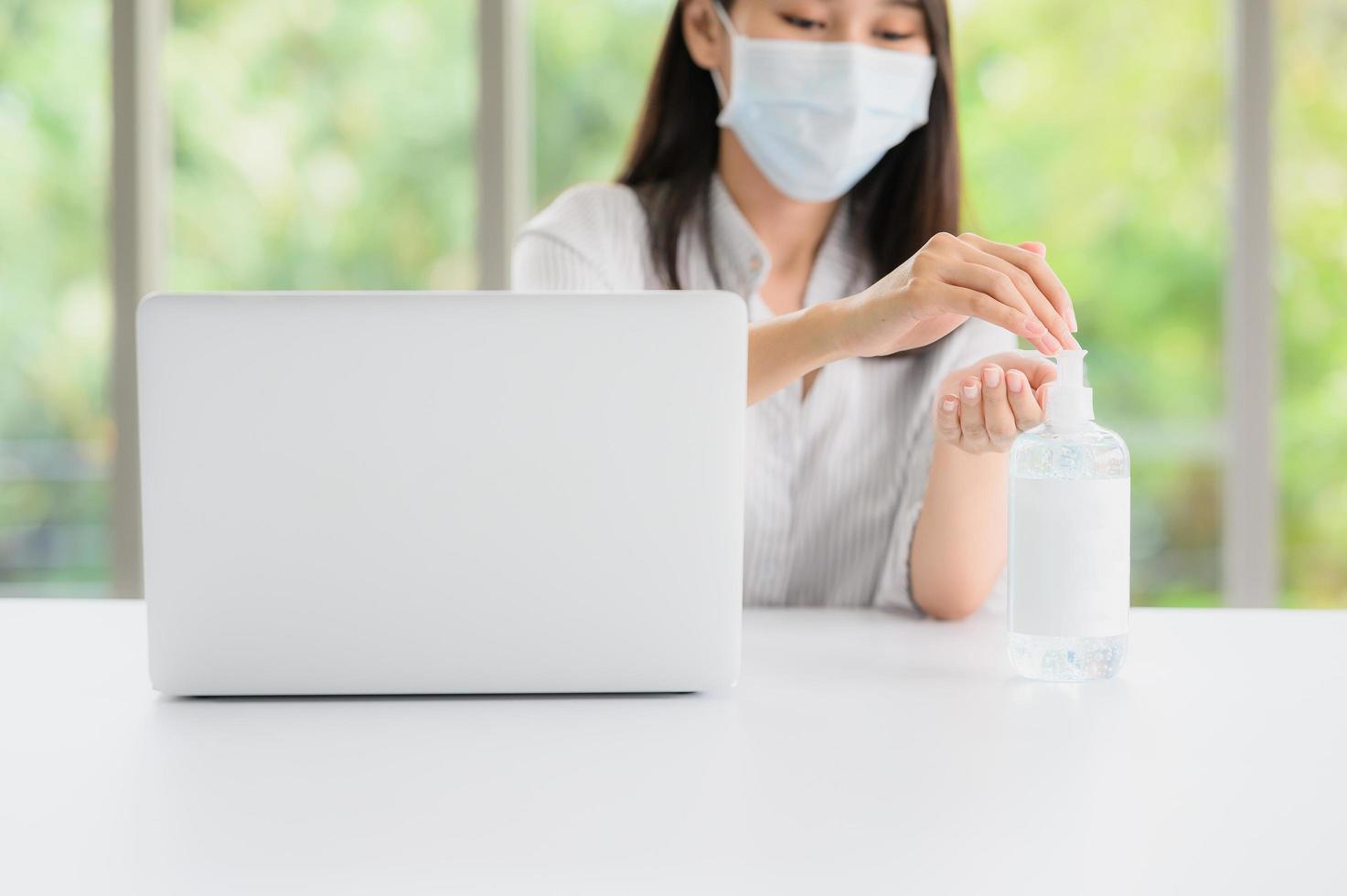 Frau, die eine Gesichtsmaske mit Desinfektionsmittel neben Computer trägt foto