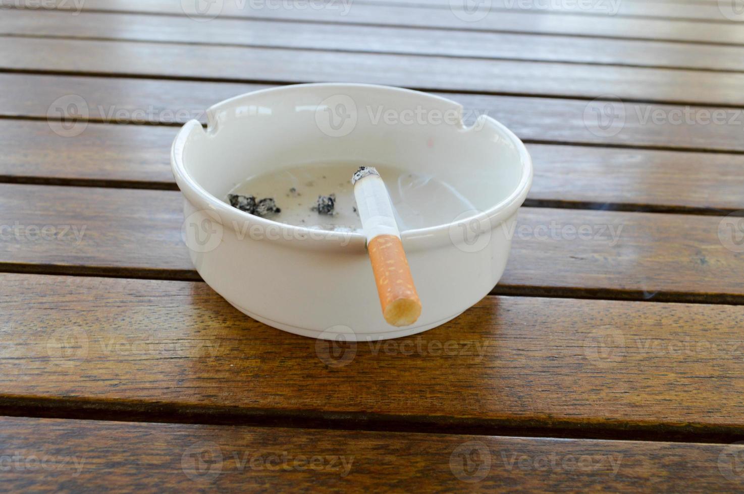 Auf einem Holztisch in einem Café steht ein weißer Keramikaschenbecher mit einer Zigarette und Asche mit einem schwarzen Feuerzeug zum Rauchen foto