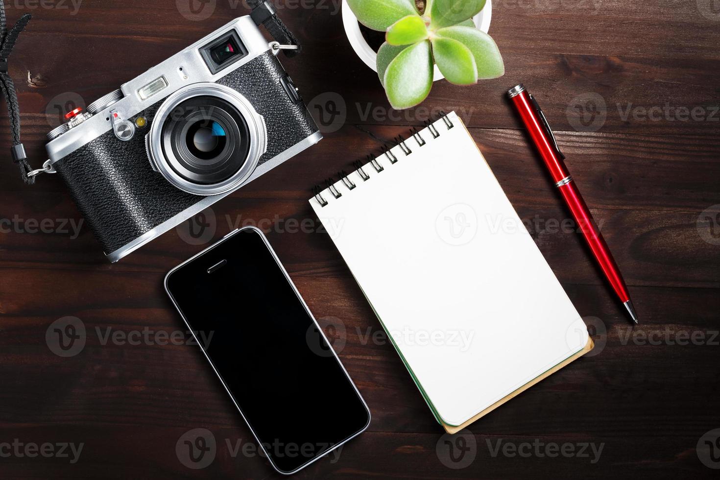 Klassische Kamera mit leerer Notizblockseite und rotem Stift auf dunkelbraunem Holztisch, Vintage-Tisch mit Telefon und grüner Blume foto