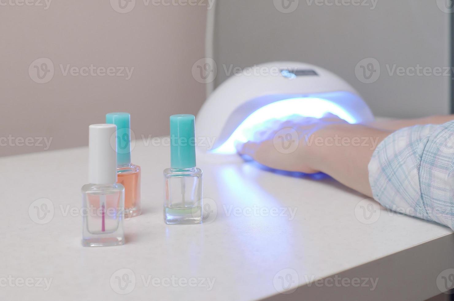 drei farblose transparente nagellackflaschen im hintergrund weiblicher hände in gel-uv-led-nagelweißlampe zum trocknen der maniküre foto