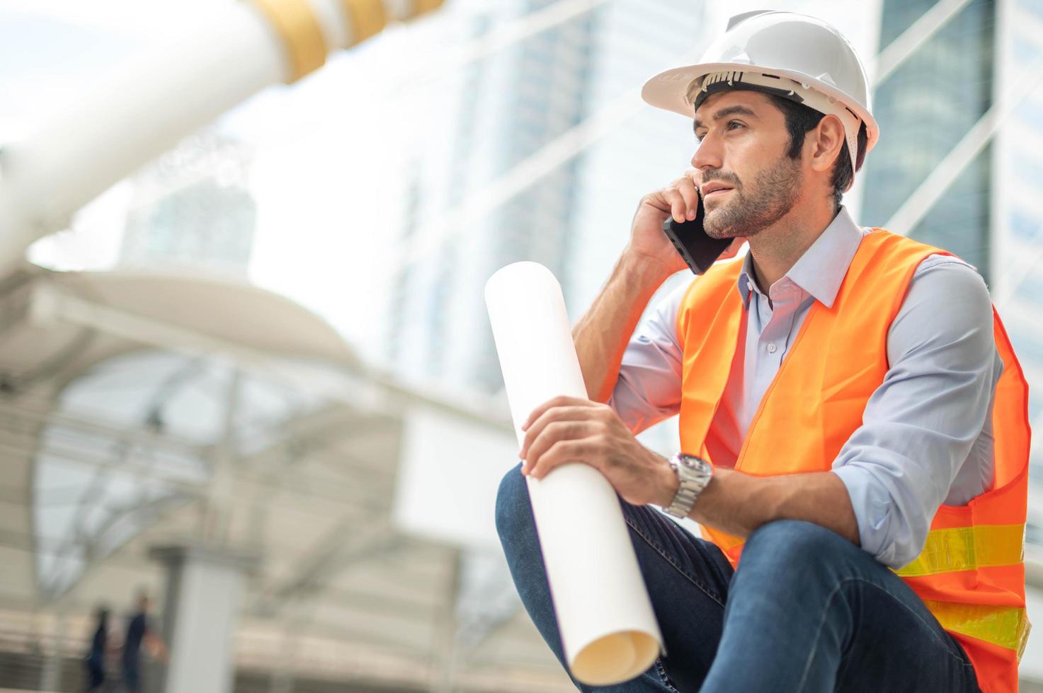 Kaukasischer Ingenieur benutzt ein Smartphone zum Reden, trägt eine orangefarbene Weste und einen großen Schutzhelm und hält in der anderen Hand den weißen Grundriss in der Baustelle der Innenstadt. foto
