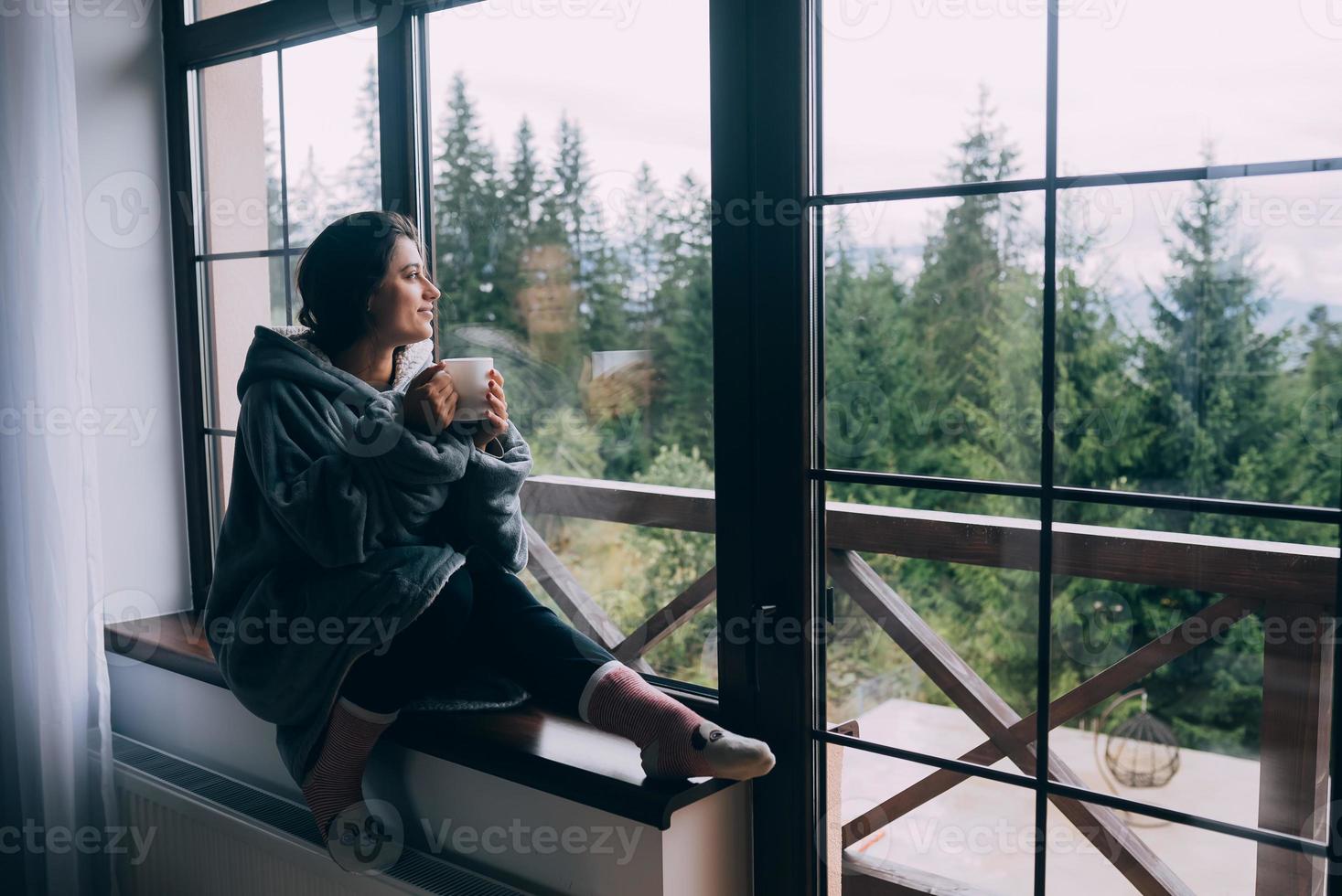 Frau hält Becher, sitzt auf der Fensterbank und blickt auf die Berge foto