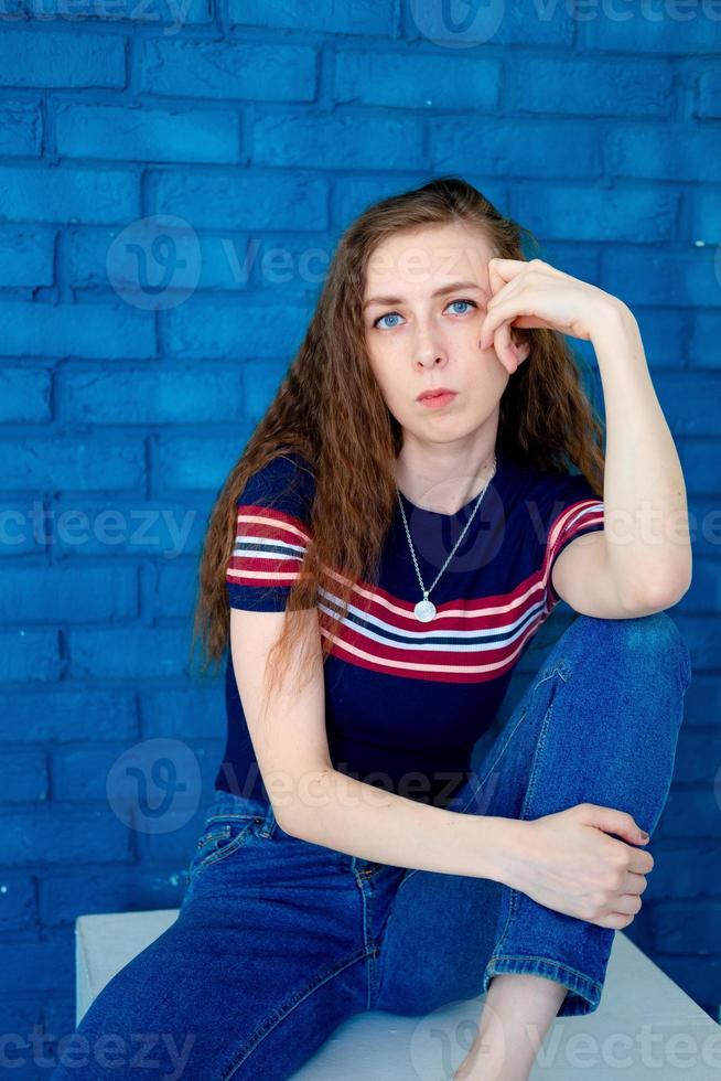 Porträt eines jungen und ernsthaften Mädchens, das auf einem Stuhl sitzt, auf einem blauen Backsteinhintergrund, mit langen Haaren, einem engen T-Shirt und Jeans foto