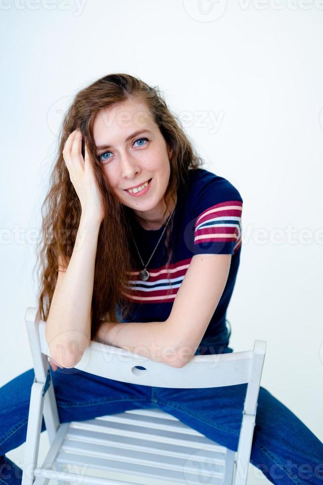 Porträt eines jungen lächelnden Mädchens, das auf einem Stuhl sitzt, auf weißem Hintergrund, mit langen Haaren, in einem engen T-Shirt und Jeans foto