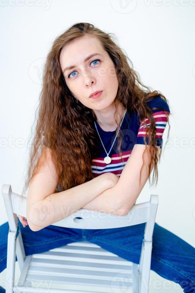 Porträt eines jungen und ernsthaften Mädchens, das auf einem Stuhl sitzt, auf weißem Hintergrund, mit langen Haaren, einem engen T-Shirt und Jeans foto