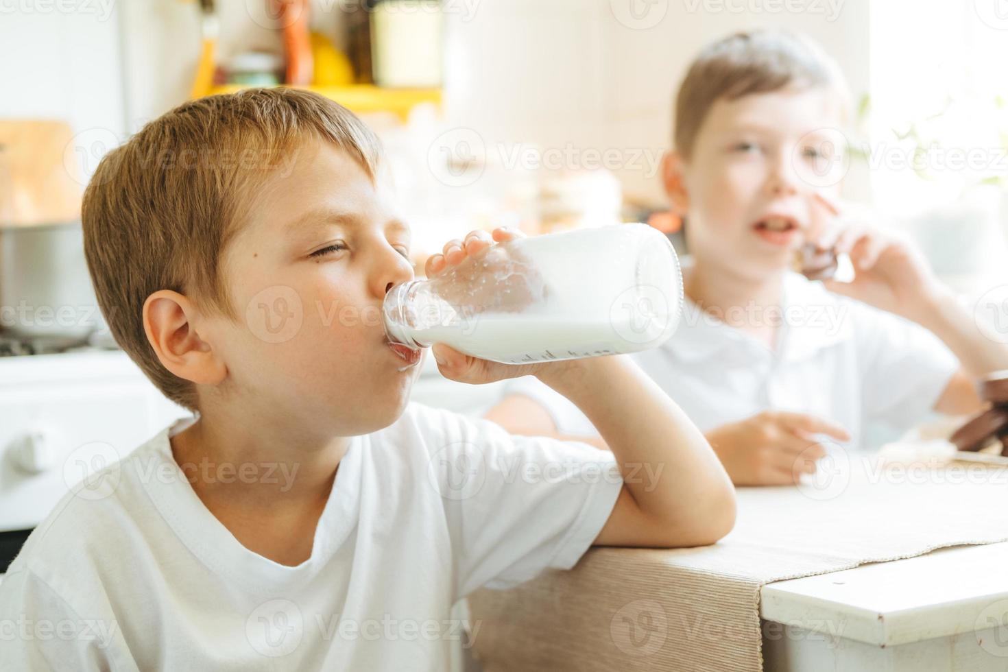 ein junge trinkt zu hause milch aus einer flasche in der küche. Morgenfrühstück mit Milch. glückliches Kind in einem weißen T-Shirt trinkt Milch aus einer Flasche foto