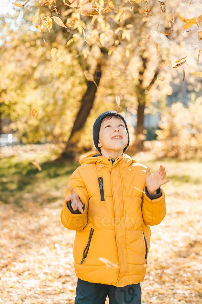 junge in einer gelben jacke, streut blätter in einem herbstpark. das kind freut sich über die herbstblätter. glückliche Kindheit. leuchtend gelbe Jacke und Blätter foto