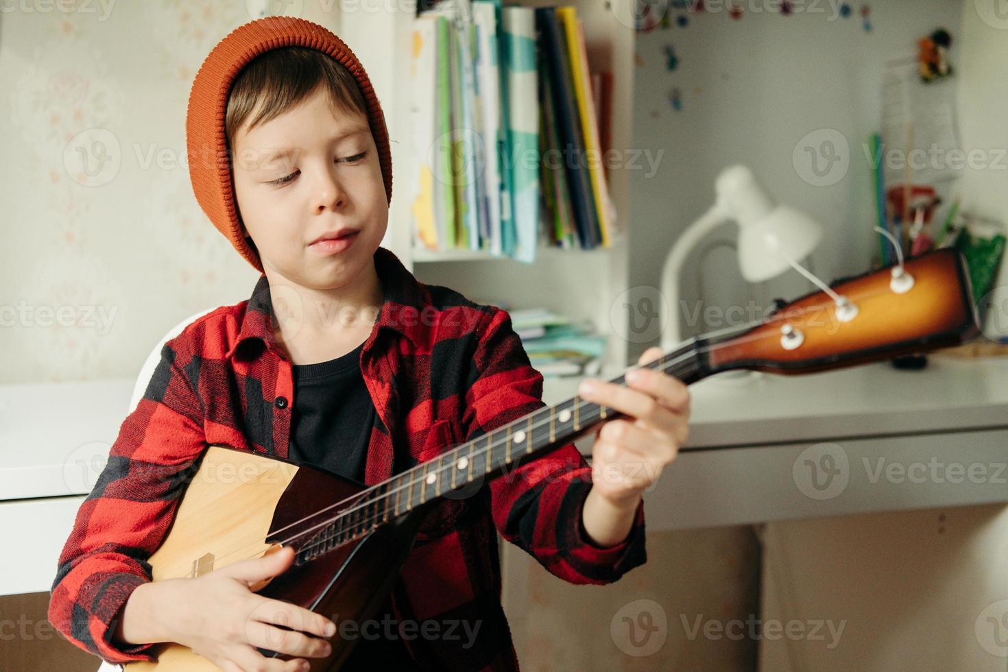 Junge mit rotem Hut und kariertem Hemd spielt Balalaika. hübscher junge, der seine gitarre hält. Musikunterricht zu Hause. Hobby für die Seele. Musikunterricht zu Hause foto