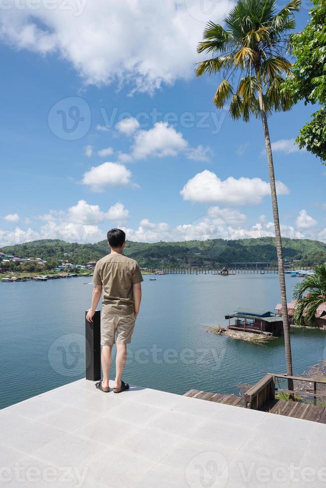 junger asiatischer mann stand an sonnigen tagen auf der terrasse und dem traditionellen dorf am seeblick foto