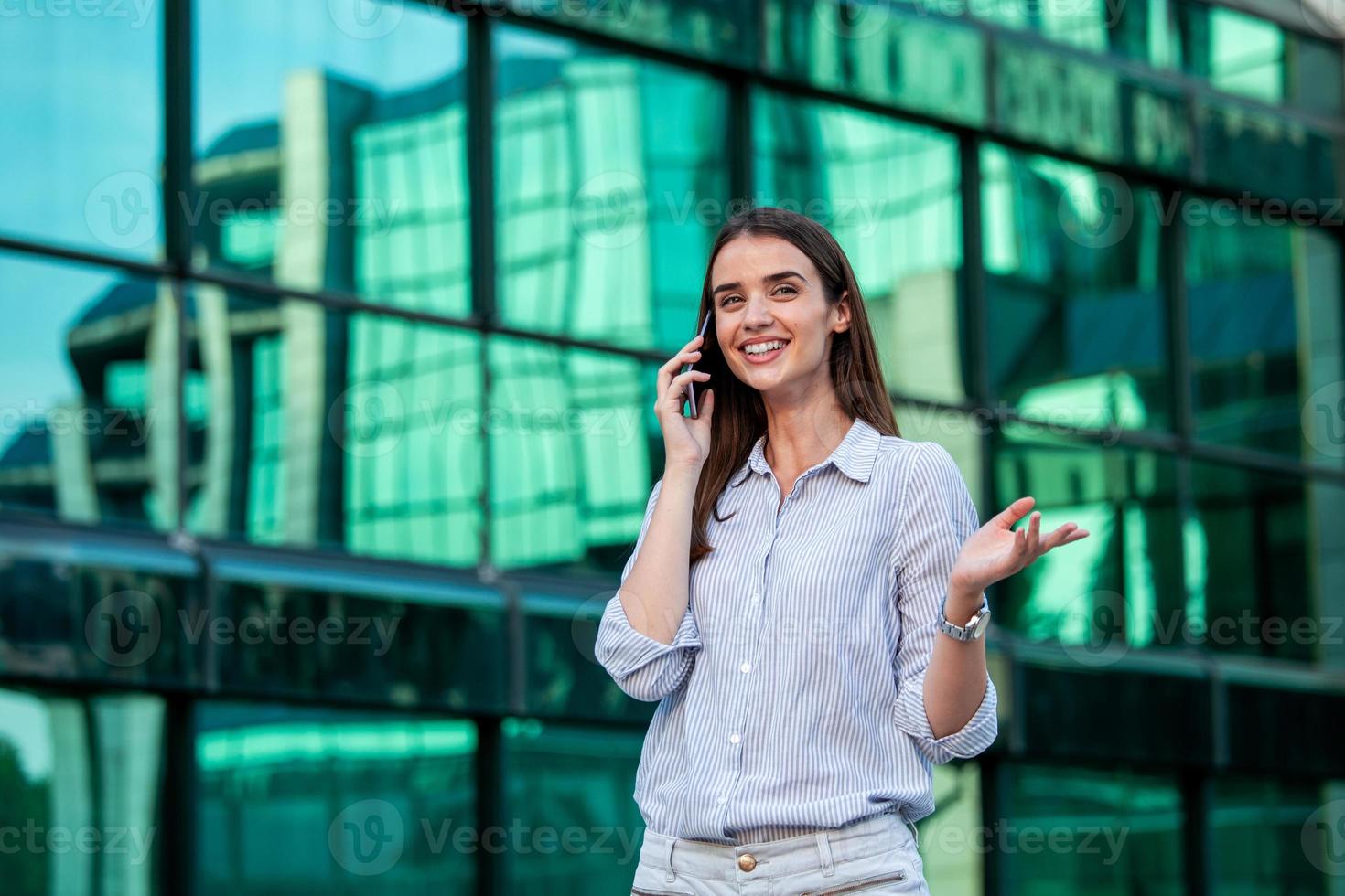 Geschäftsfrau, die mit einem mobilen Smartphone auf der Straße mit Bürogebäuden im Hintergrund spricht. junge frau mit dem smartphone, das gegen straße steht, verschwommenes gebäudehintergrund. foto