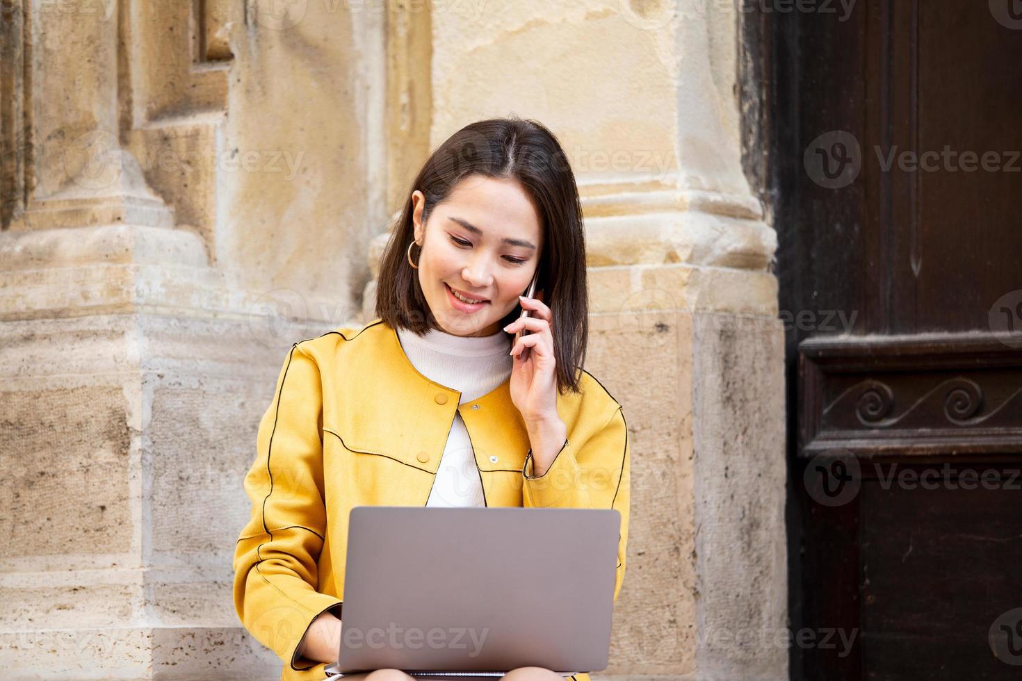 Lächelnde asiatische Frau, die an einem Laptop arbeitet, während sie auf dem Boden sitzt und mit ihrem Smartphone spricht. asiatische Frau, die auf einem Mobiltelefon spricht, während sie einen Computer-Laptop verwendet foto