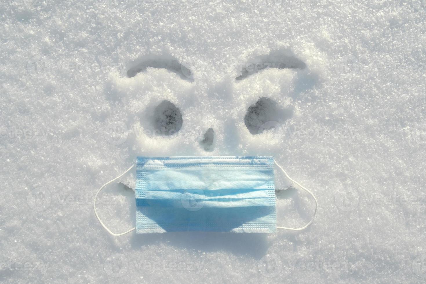 ein aufgeregter, in den Schnee gemalter Smiley mit einer echten medizinischen Maske im Gesicht. Gesundheitskonzept. foto