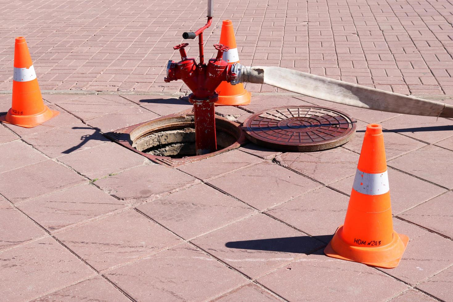 Ein großer Hydrant aus rotem Metall mit Schläuchen und Manschetten, die mit der Wasserquelle in der Kanalluke verbunden sind, und dreieckigen orangefarbenen Notkegeln, die vor der Gefahr warnen foto