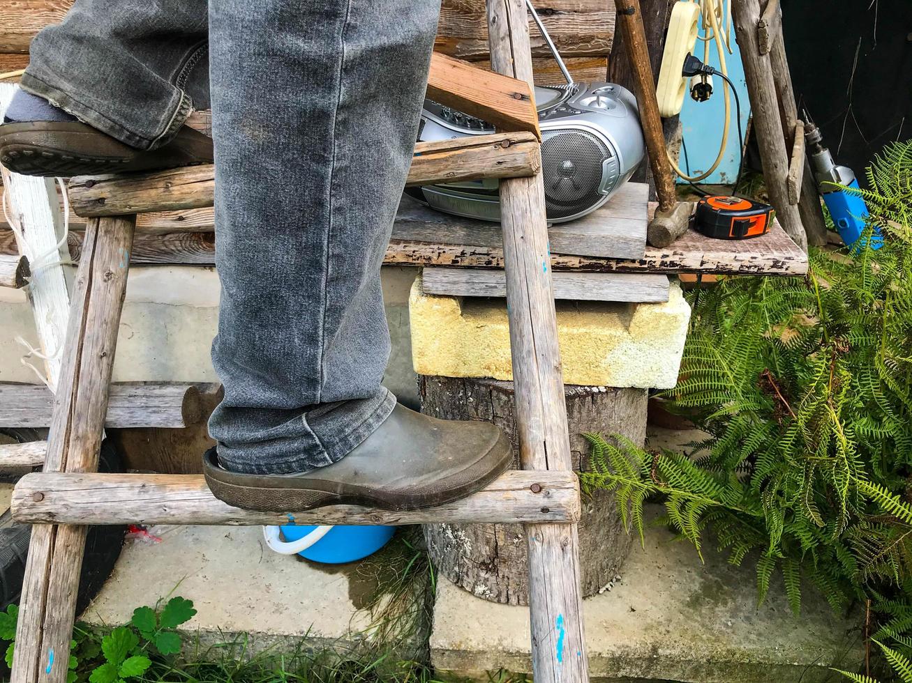 ein mann steht in schwarzen gummigaloschen auf einer holzleiter. Arbeit in der Höhe. schmutzige Schuhe, Landwirtschaft. vor dem Hintergrund einer Holzbank, Tonbandgerät, Farnblätter foto