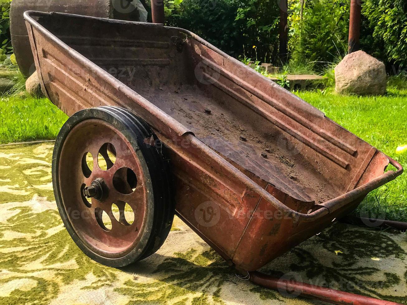 Auf einem Teppich steht eine Schubkarre aus Metall mit großen Rädern. Gartenpflege in Russland. Überall Teppiche, Mode aus den 90ern. reiches Interieur, Dekoration der Natur und Sommerhäuser foto