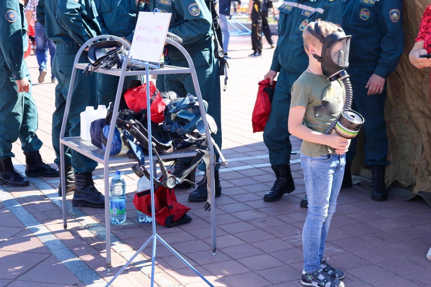 Ein kleines Kind trägt eine Schutzmaske in einer Gasmaske und hält eine Schachtel Gasmasken in der Umgebung von Feuerwehrleuten und Militärs. weißrussland, minsk, 08.08.2018 foto