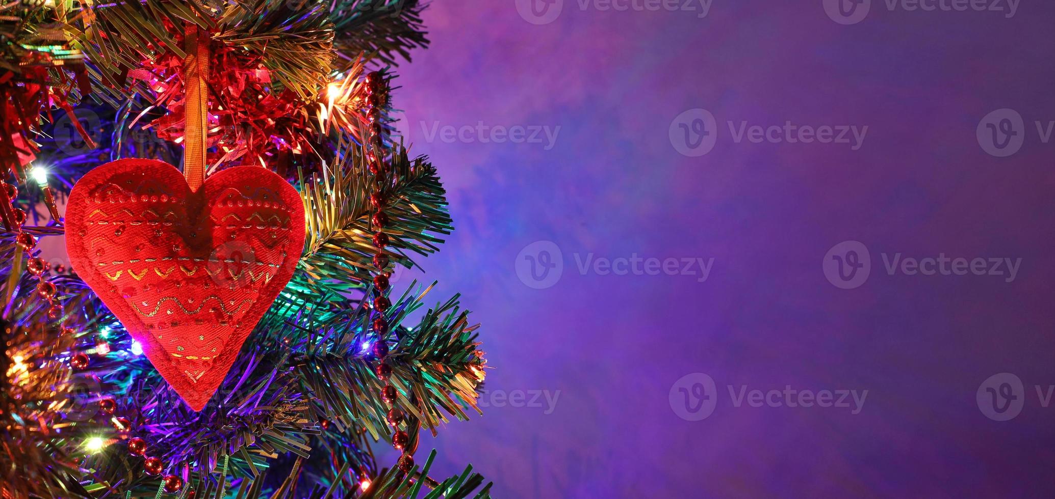 Rotes Herz, handgefertigte Weihnachtsdekoration mit Ornament auf Tanne in der Feiertagsnacht, bunte Bokeh-Girlande, rote Perlen, unscharfer dunkelvioletter Hintergrund. weihnachtsbanner mit kopierraum. foto