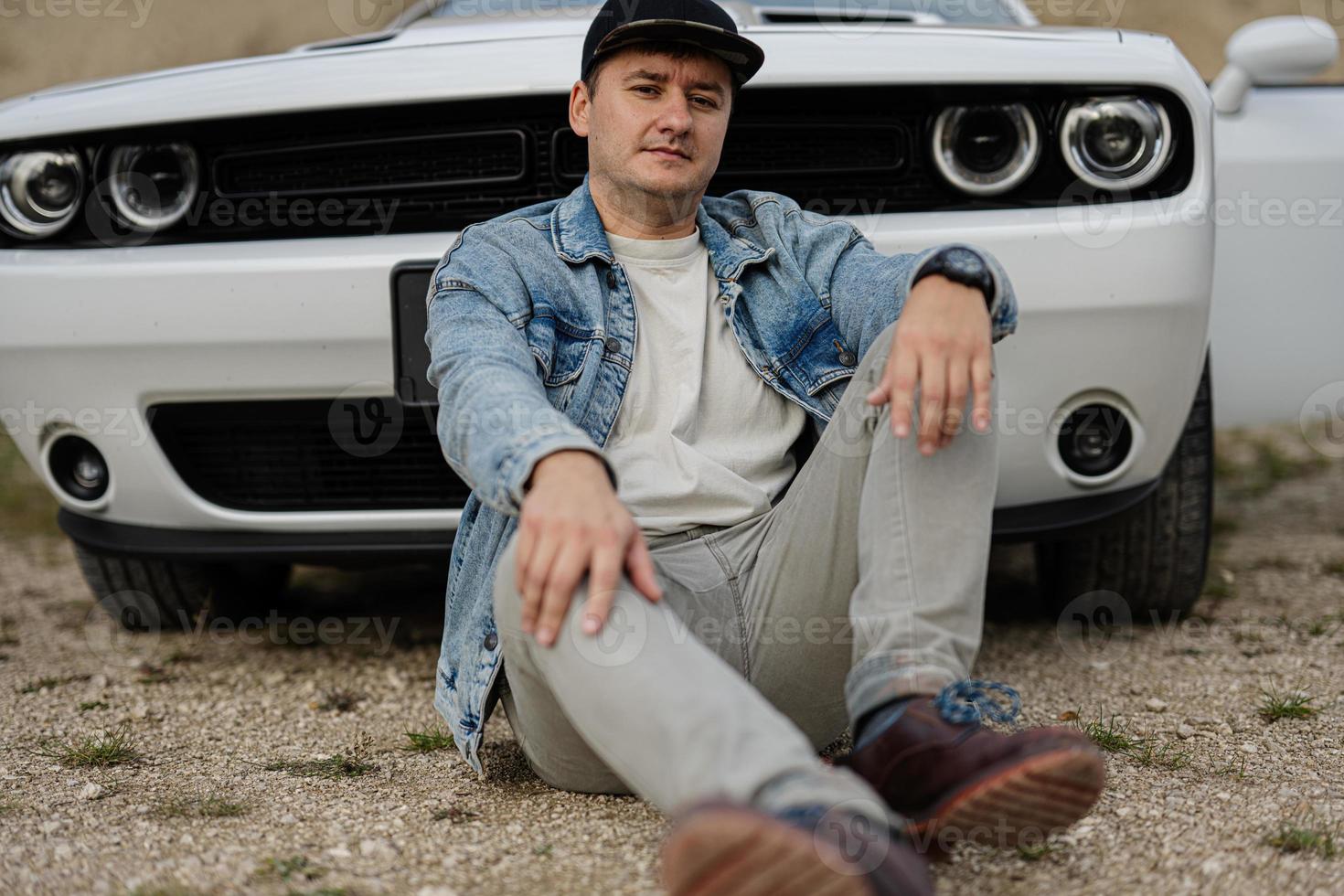 Gutaussehender Mann in Jeansjacke und Mütze, der in der Karriere neben seinem weißen Muscle-Car sitzt. foto