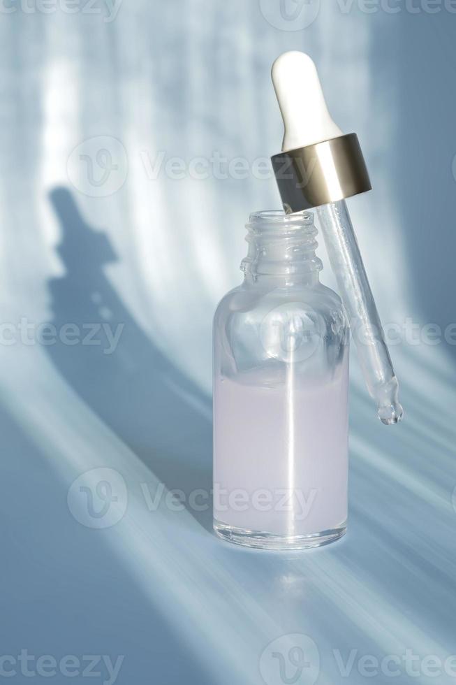 eine tropfflasche aus glas mit schönheitspflegeserum, hyaluronsäure und vitaminen auf blauem hintergrund mit trendigen harten schatten. heimkosmetik und spa-konzept foto