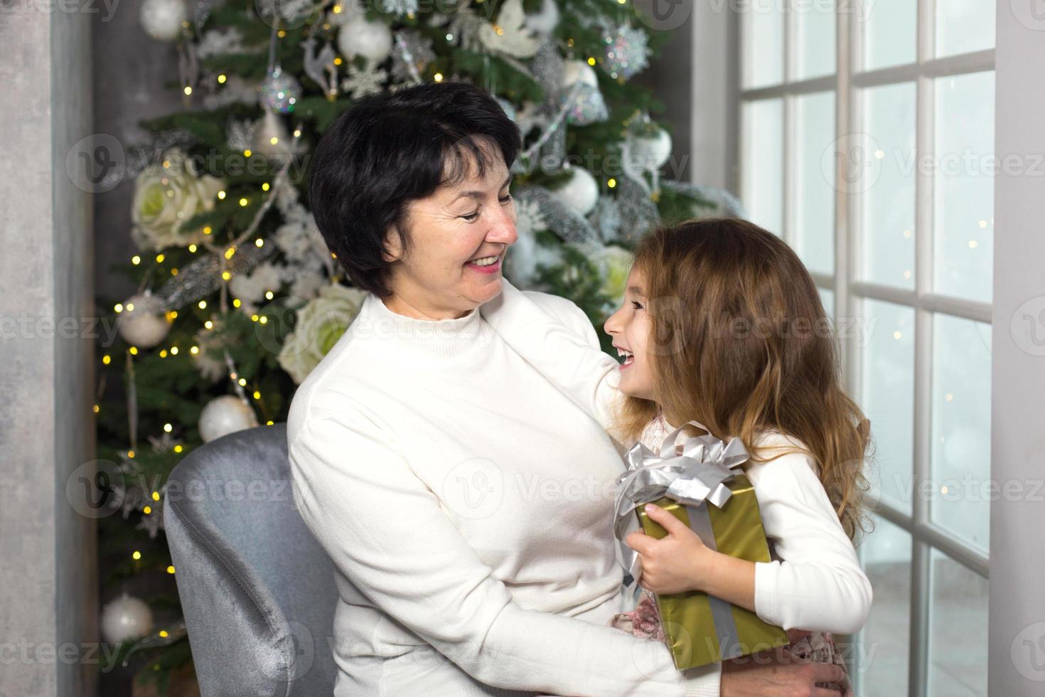 großmutter mit einem kleinen mädchen auf dem hintergrund von weihnachtsdekorationen und einem großen fenster. familienurlaub, emotionen, geschenkbox. Enkelin auf Omas Schoß. Neujahr foto