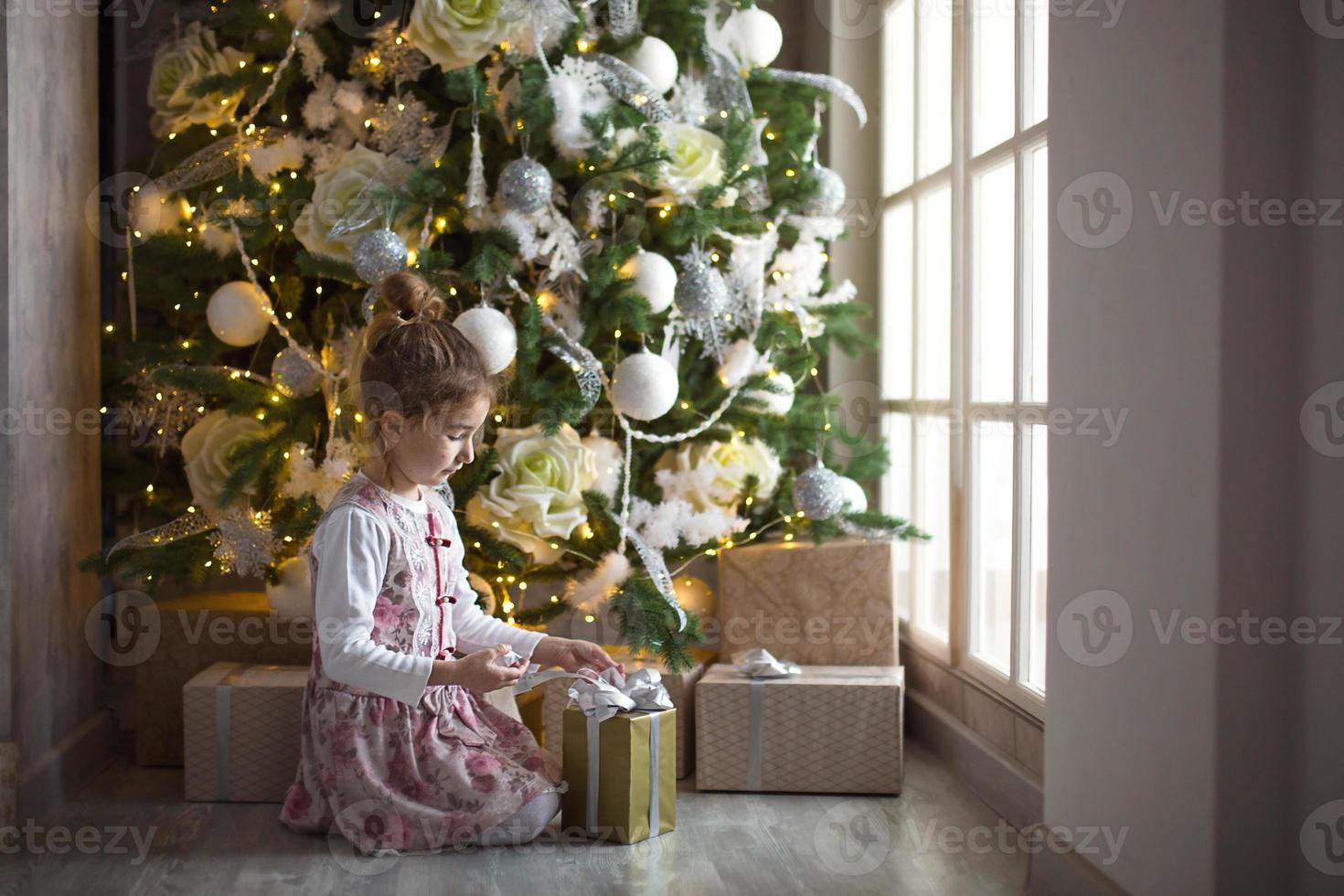 Kleines Mädchen in schönem Kleid sitzt unter dem Weihnachtsbaum mit Geschenkbox und Schleife. licht aus einem großen fenster, dunkler raum, wohnkomfort, weihnachten, magie und freude. neujahr, weißes dekor foto