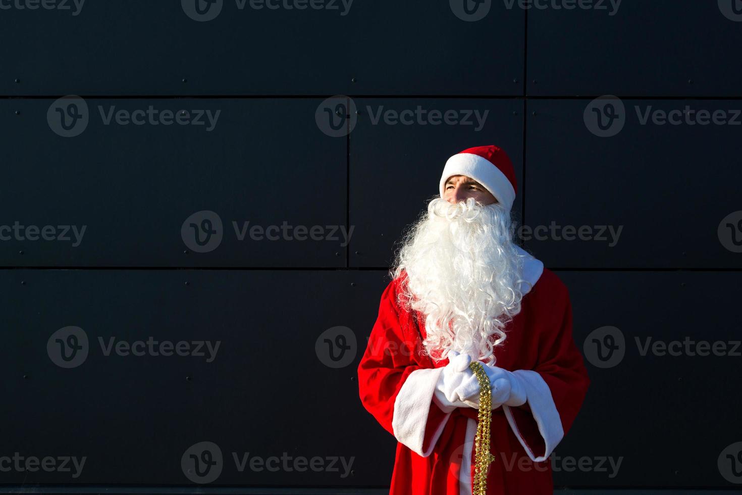 ein junger mann, der als weihnachtsmann auf der straße verkleidet ist und an einer grauen modernen wand steht. weißer Bart, ein roter Anzug mit Hut. weihnachten, neujahr. Russischer Großvater Frost. grauer moderner hintergrund, kopierräume foto