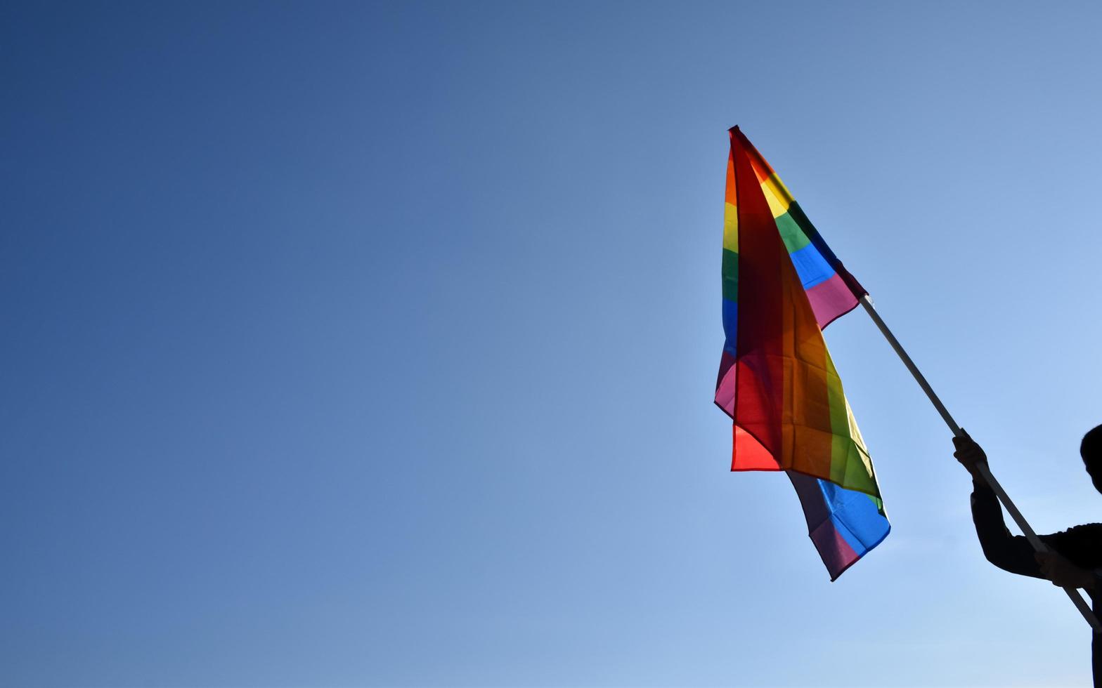 regenbogenfahne, die in der hand vor blauem hintergrund hält, konzept für die lgbt-feier im stolzmonat juni auf der ganzen welt. foto