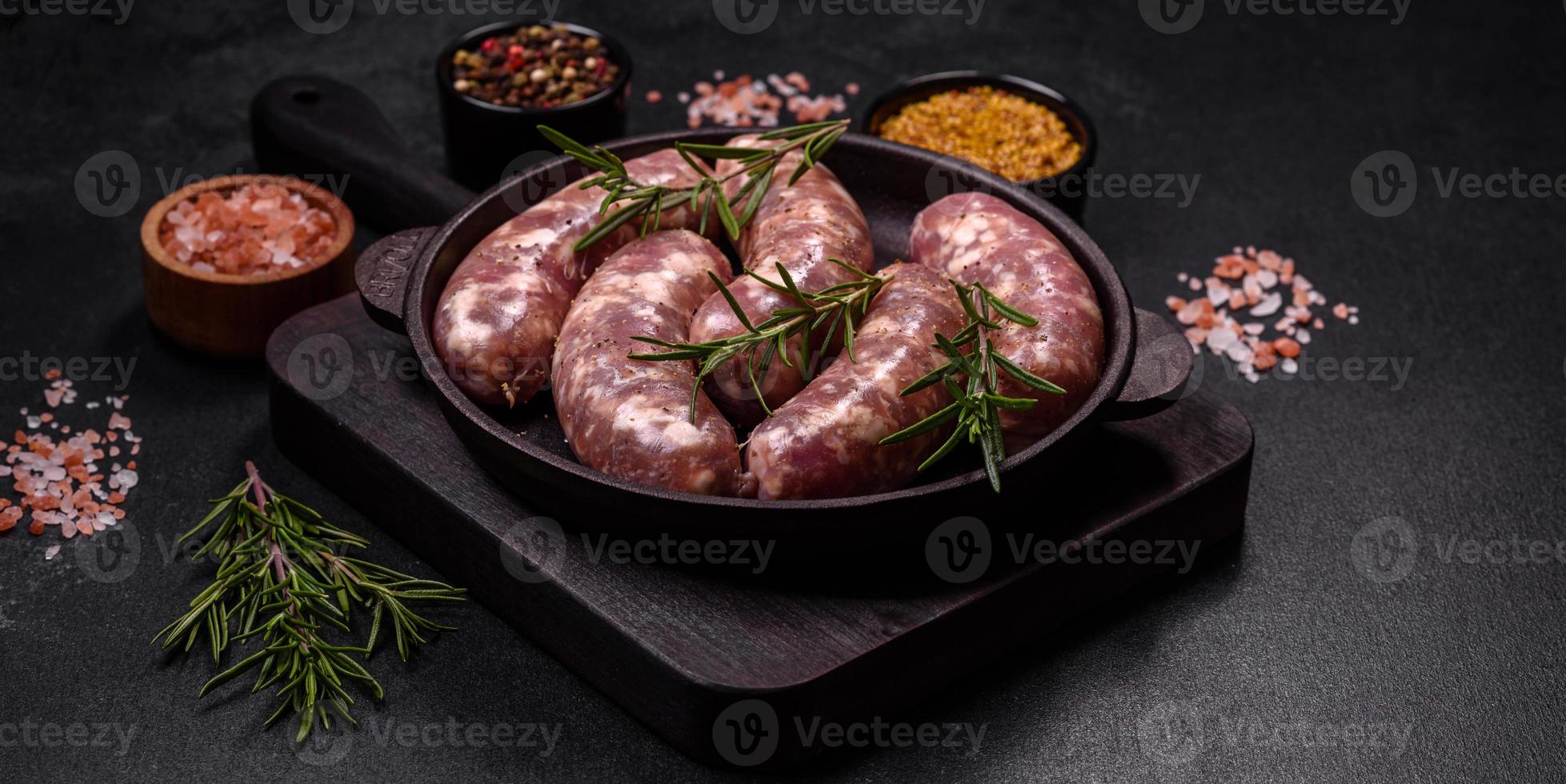 Rohe Schweinswürste grillen mit Gewürzen und Kräutern auf einem dunklen Betontisch foto