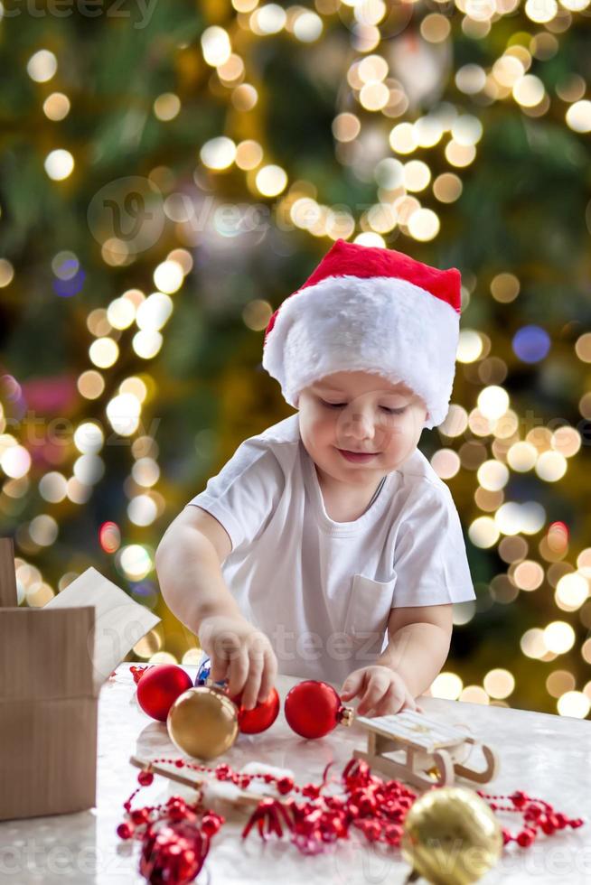 Kind Junge nimmt Weihnachtsspielzeug aus der Schachtel, um den Baum zu verkleiden. der weihnachtsbaum wird beleuchtet und das baby betrachtet die weihnachtsspielzeuge mit vergnügen und freudigen gefühlen. foto