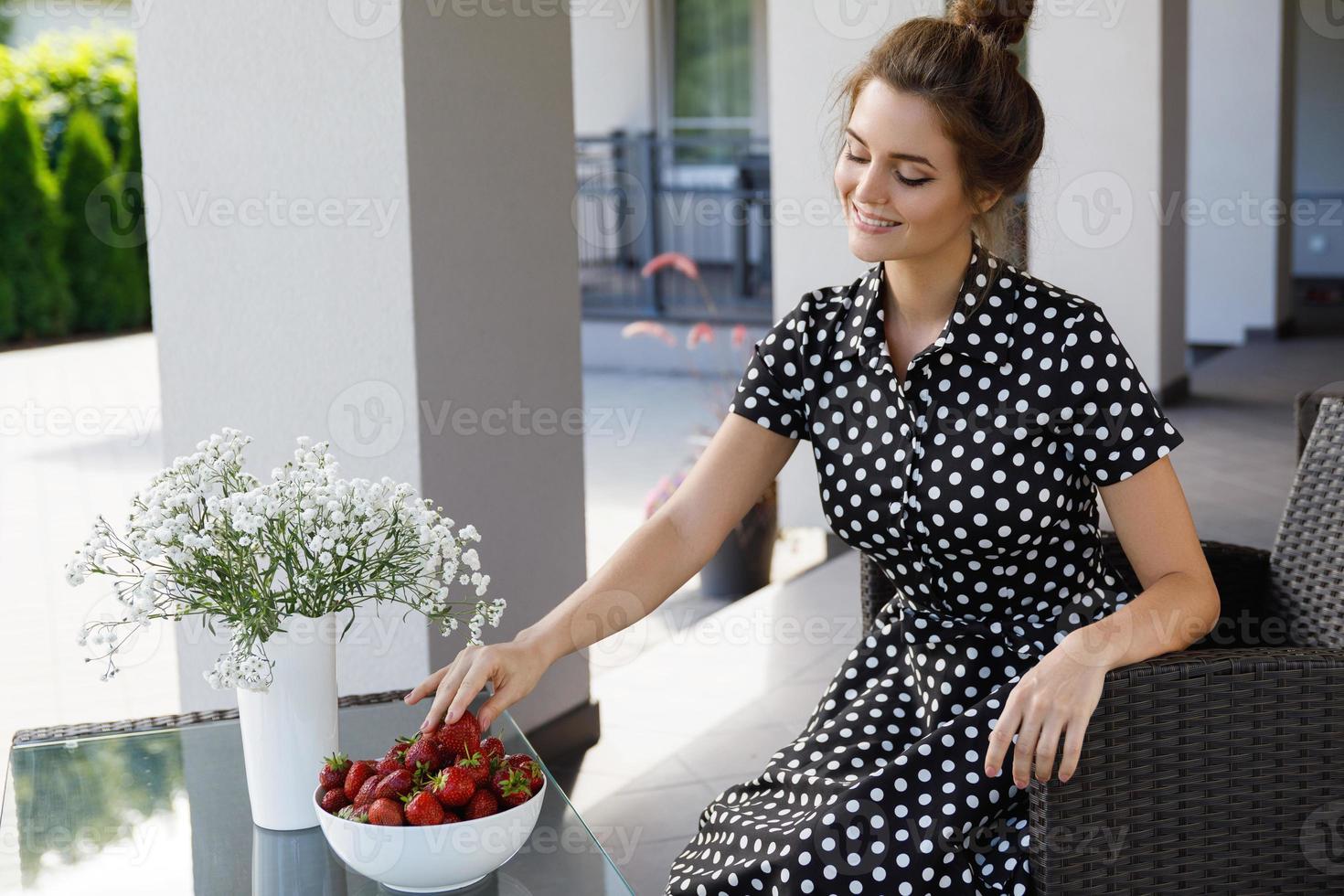 Wunderschöne Frau, die ein wunderschönes Kleid mit einem Tupfenmuster trägt, das auf einer Terrasse sitzt und Erdbeeren isst foto