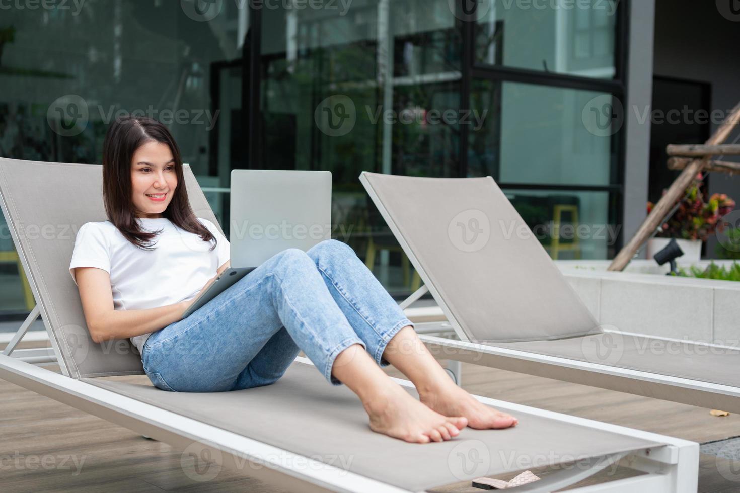 Fröhliche junge Unternehmerin, die auf einem Solarium neben dem Pool sitzt und einen Laptop für Remote-Online-Arbeiten verwendet, digitales Online-Geschäftsprojekt im ruhigen Hof des Resorthauses, Arbeit im Urlaub foto