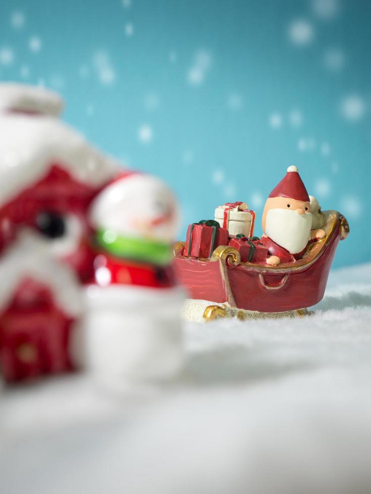 glücklicher weihnachtsmann mit geschenkbox auf dem schneeschlitten, der zum haus geht. in der nähe des hauses haben schneemann und weihnachtsbaum. weihnachtsmann und haus im schnee. weihnachts- und frohes neues jahrkonzept. foto