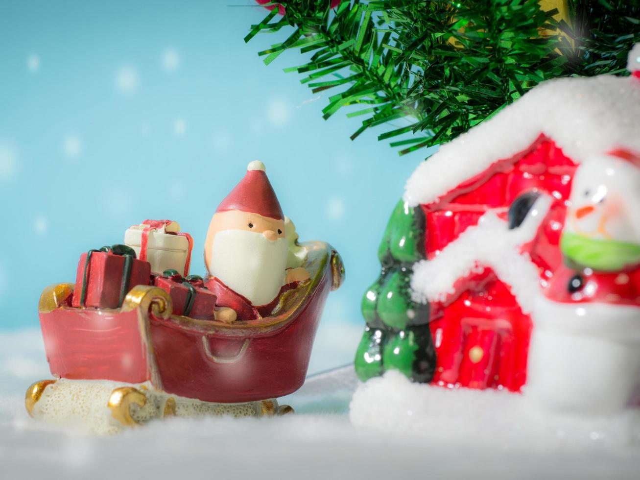 glücklicher weihnachtsmann mit geschenkbox auf dem schneeschlitten, der zum haus geht. in der nähe des hauses haben schneemann und weihnachtsbaum. weihnachtsmann und haus im schnee. weihnachts- und frohes neues jahrkonzept. foto