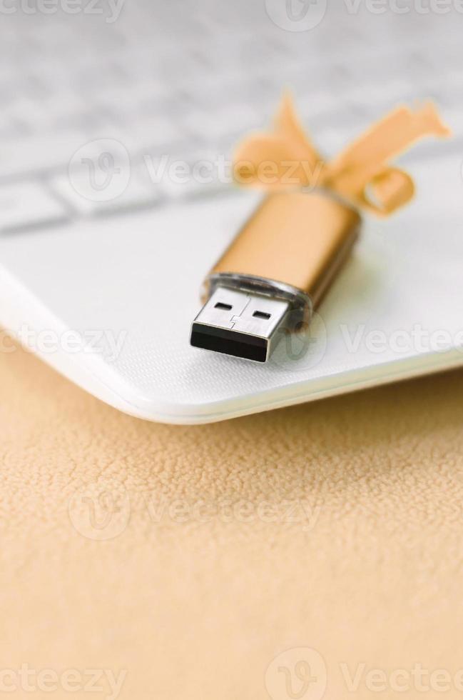 Eine orangefarbene USB-Flash-Speicherkarte mit einer Schleife liegt auf einer Decke aus weichem und pelzigem, hellorangefarbenem Fleece-Stoff neben einem weißen Laptop. klassisches weibliches Geschenkdesign für eine Speicherkarte foto