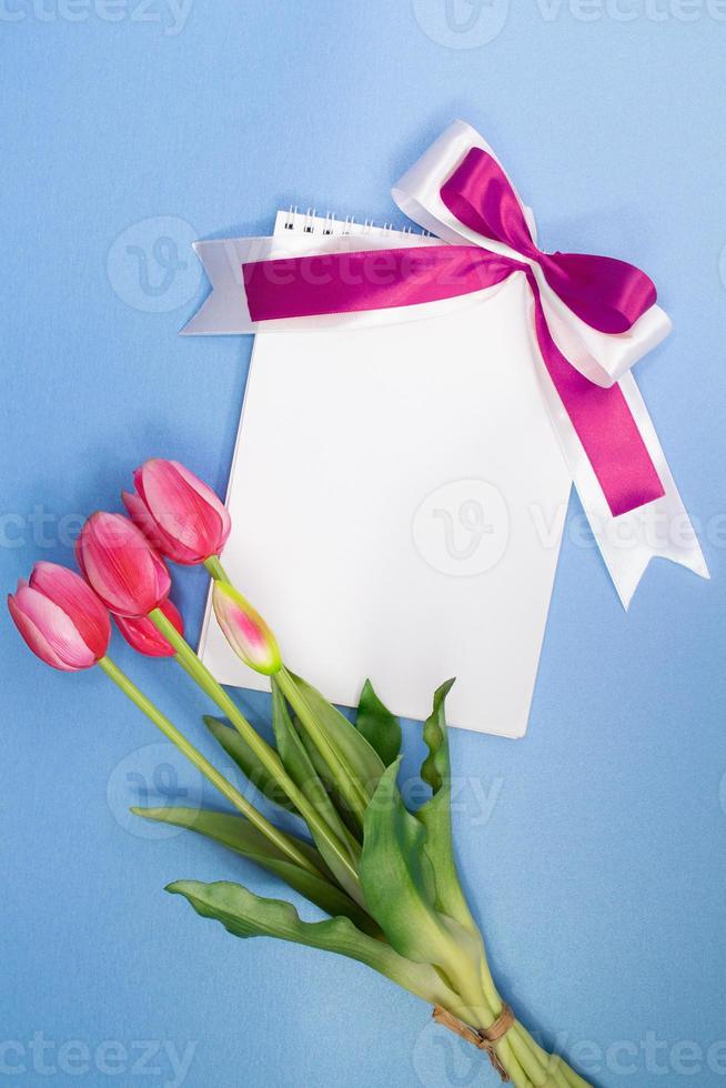 Blumenstrauß aus rosa Tulpen und Papier für Text mit lila Schleife auf blauem Hintergrund. feiertag, valentinstag, internationaler frauen- und muttertag, 8. märz, geburtstag. Platz kopieren foto