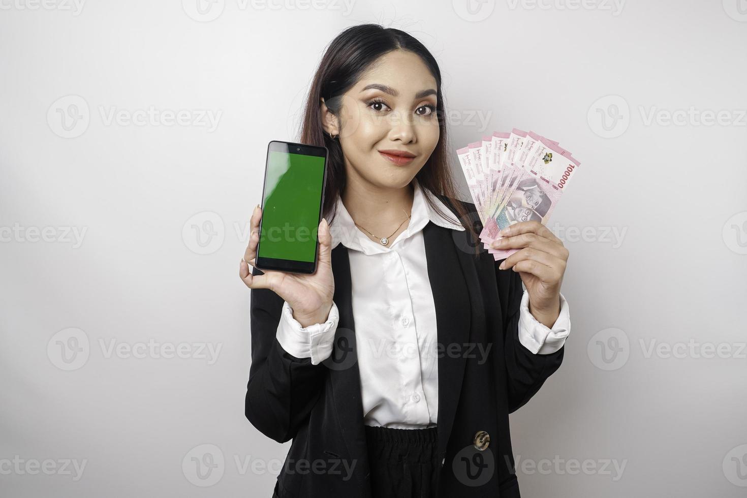 eine glückliche junge geschäftsfrau trägt einen schwarzen anzug und zeigt ihr telefon und geld in indonesischen rupiah isoliert durch weißen hintergrund foto